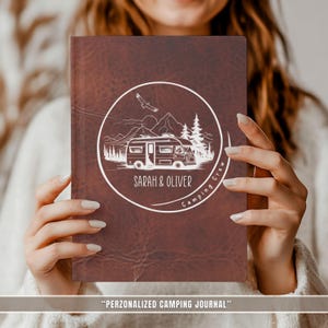 Könnte beinhalten: Ein braunes Lederjournal mit einer weißen kreisförmigen Grafik, die einen Wohnwagen, Berge, Bäume und den Text "SARAH & OLIVER" zeigt. Das Journal trägt auch den Text "Camping Crew" und "PERZONALIZED CAMPING JOURNAL".