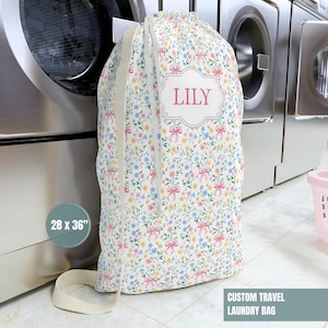 May include: A floral print laundry bag with a drawstring closure. The bag features a pattern of small flowers and bows in pink, blue, and yellow on a white background. The bag is 28 x 36 inches and has the name "LILY" on it.