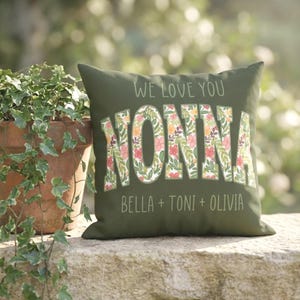 May include: Olive green decorative pillow with the text "WE LOVE YOU NONNA" in white. The word "NONNA" is filled with a floral pattern of pink, orange, and green. The names "BELLA + TONI + OLIVIA" are printed below.