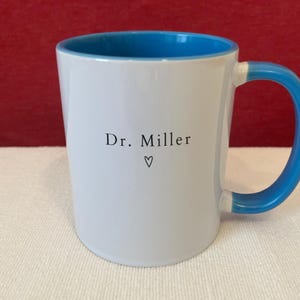 May include: A white ceramic mug with a bright blue interior and a matching blue handle. The exterior features the black printed text Dr. Miller centered on the front, positioned above a small, simple black outline of a heart symbol.