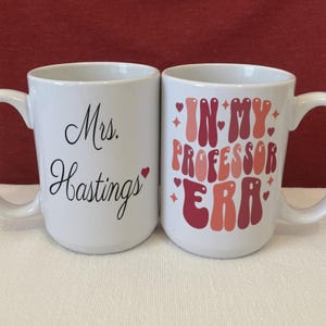 May include: Two white ceramic mugs. The left mug features the black text "Mrs. Hastings" with a small red heart. The right mug displays the red and pink text "IN MY PROFESSOR ERA" in a retro font, surrounded by small red hearts and star icons.