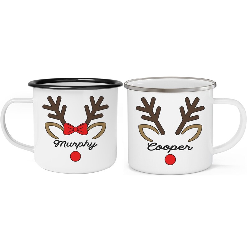 Puede incluir: Dos tazas de esmalte blanco con dise&ntilde;os de cara de reno. Una taza tiene un borde negro, un lazo rojo y el nombre "Murphy". La otra tiene un borde plateado y el nombre "Cooper". Ambas tazas tienen cuernos marrones, orejas beige y una nariz roja.