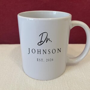 May include: A white ceramic mug featuring black printed text that reads Dr. JOHNSON EST. 2026. The mug has a smooth finish, a cylindrical body, and a curved handle on the right side, providing a clean and simple design for holding hot beverages.
