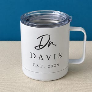 May include: A white stainless steel mug with a clear plastic lid and a side handle. The front features black printed text that reads Dr. DAVIS EST. 2026, with the title in a script font and the name and year in a clean, capitalized serif typeface.