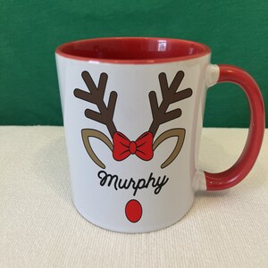 Puede incluir: Taza de cer&aacute;mica blanca con interior y asa rojos. La taza presenta un dise&ntilde;o de cara de reno con cuernos marrones, una pajarita roja y una nariz roja. El nombre "Murphy" est&aacute; escrito debajo.