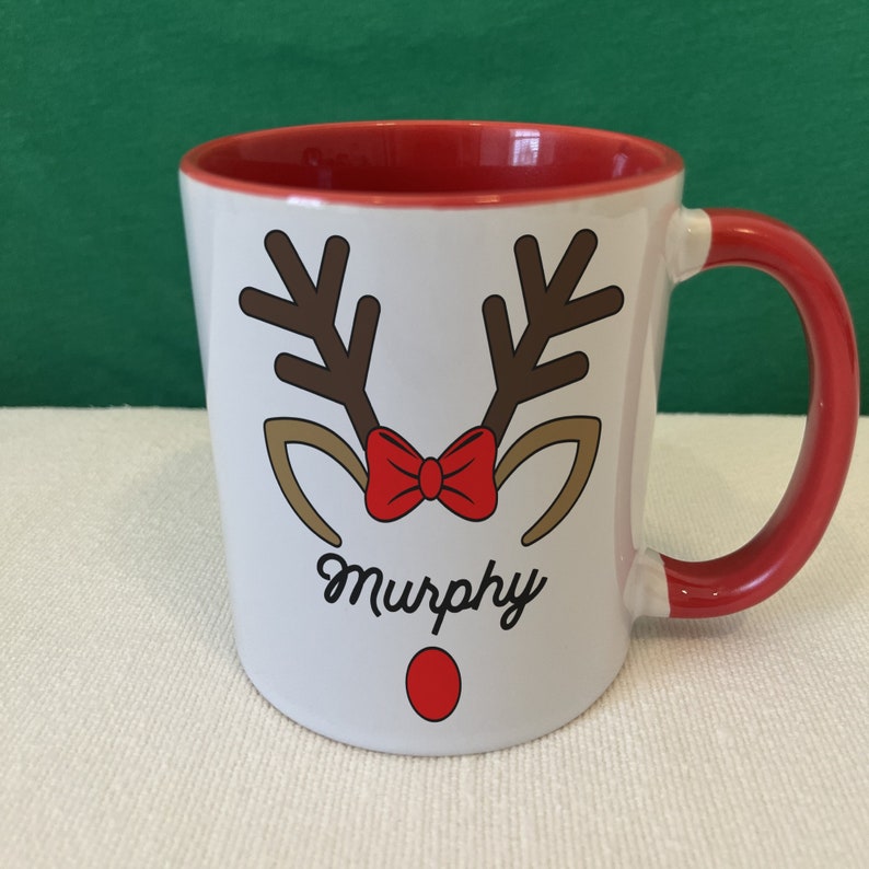 Puede incluir: Taza de cer&aacute;mica blanca con interior y asa rojos. La taza presenta un dise&ntilde;o de cara de reno con cuernos marrones, una pajarita roja y una nariz roja. El nombre "Murphy" est&aacute; escrito debajo.