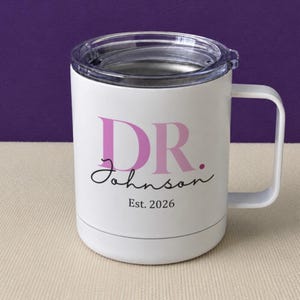 May include: A white stainless steel mug with a clear plastic lid and a side handle. The front features the pink text DR., the black cursive text Johnson, and the black text Est. 2026 printed in a clean, centered layout on the smooth exterior surface.