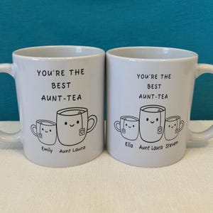 May include: Two white ceramic mugs featuring black line art of smiling teacups with tea bags. The left mug says YOU'RE THE BEST AUNT-TEA with names Emily and Aunt Laura. The right mug says the same with names Ella, Aunt Laura, and Steven.