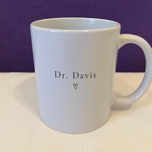 May include: A white ceramic mug featuring the black printed text Dr. Davis centered on the front. Below the name is a small, simple black outline of a heart. The mug has a smooth, glossy finish and a curved handle on the right side of the body.