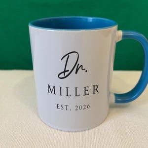 May include: A white ceramic mug with a bright blue interior and handle. The front features black text that reads Dr. MILLER EST. 2026, with the title in a cursive script font and the name and date in a clean, capitalized sans-serif typeface.