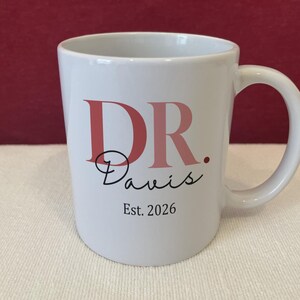 May include: A white ceramic mug featuring the text DR. in large pink letters, followed by the name Davis in a black cursive script, and the text Est. 2026 in a simple black serif font centered below the name on the front of the mug.