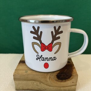 Puede incluir: Taza de esmalte blanco con dise&ntilde;o de cara de reno. La taza presenta cuernos marrones, una pajarita roja y el nombre "Hanna" en fuente cursiva. Un punto rojo sirve como nariz del reno. La taza tiene un borde plateado y un asa blanca.