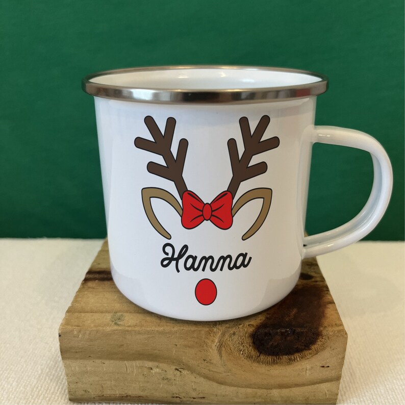 Puede incluir: Taza de esmalte blanco con dise&ntilde;o de cara de reno. La taza presenta cuernos marrones, una pajarita roja y el nombre "Hanna" en fuente cursiva. Un punto rojo sirve como nariz del reno. La taza tiene un borde plateado y un asa blanca.
