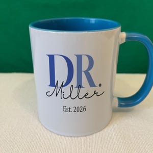 May include: A white ceramic mug with a bright blue interior and handle. The front features the text DR. in blue, followed by the name Miller in black cursive script, and the text Est. 2026 in black print centered below the name.