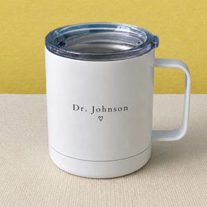 May include: A white insulated mug with a clear plastic lid and a side handle. The front of the mug features the black printed text Dr. Johnson with a small heart symbol centered directly underneath the name in a simple, clean font style.