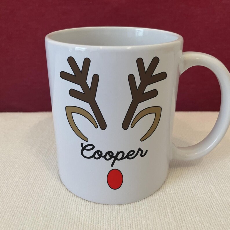 Puede incluir: Taza de cer&aacute;mica blanca con un dise&ntilde;o de cara de reno. La taza presenta cuernos marrones, formas de ojos beige, una nariz roja y el nombre "Cooper" en escritura negra. La taza tiene un asa blanca y es probablemente para bebidas calientes.