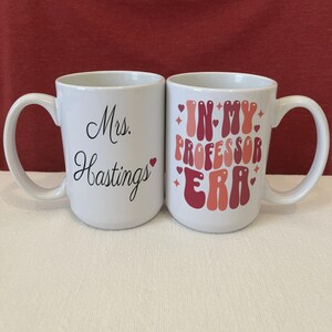 May include: Two white ceramic mugs. The left mug features the black text Mrs. Hastings with a small red heart. The right mug displays the red and pink text IN MY PROFESSOR ERA in a bold, wavy font, accented with small red hearts and star shapes.