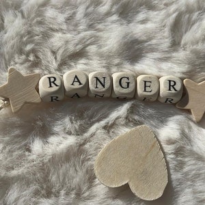 May include: Wooden beads spelling out the name "Ranger" with two wooden stars at either end. The beads are strung together with a white string.