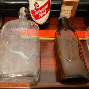 May include: Five vintage glass bottles, including a clear bottle with the text "Dixie Belle" on it, a brown bottle with a man etched into the glass, a clear bottle with a label that says "Bohemian Maid Beer", a clear bottle with a label that says "Take one to water", and a clear bottle with a label that says "Alcohol with medicinal properties".