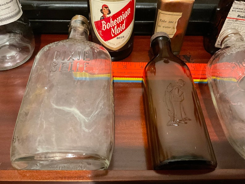 May include: Five vintage glass bottles, including a clear bottle with the text "Dixie Belle" on it, a brown bottle with a man etched into the glass, a clear bottle with a label that says "Bohemian Maid Beer", a clear bottle with a label that says "Take one to water", and a clear bottle with a label that says "Alcohol with medicinal properties".