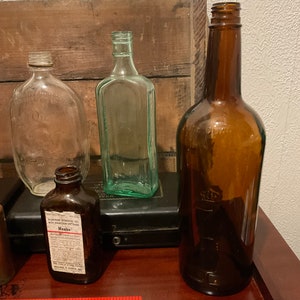 May include: Four glass bottles of various shapes and sizes. The bottles are clear, green, and brown. The bottles are sitting on a wooden surface. One bottle has a label that reads "Maalox Antacid Demulcent Concentrating".