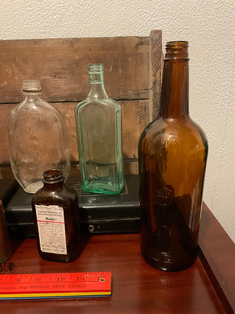 May include: Four glass bottles of various shapes and sizes. The bottles are clear, green, and brown. The bottles are sitting on a wooden surface. One bottle has a label that reads "Maalox Antacid Demulcent Concentrating".