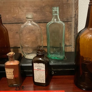 May include: A collection of vintage glass bottles, including a clear bottle with a label that reads "Maalox Antacid Demulcent Non-Constipating". The bottles are arranged on a wooden surface with a red ruler.