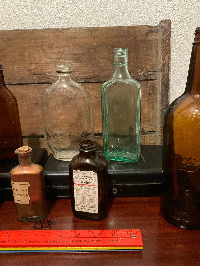May include: A collection of vintage glass bottles, including a clear bottle with a label that reads "Maalox Antacid Demulcent Non-Constipating". The bottles are arranged on a wooden surface with a red ruler.