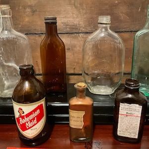 May include: A collection of vintage glass bottles, including a clear glass bottle with a cork stopper, a brown glass bottle with a label that reads "Bohemian Maid Beer", a clear glass bottle with a label that reads "Take one teaspoon in water", a brown glass bottle with a label that reads "Maalox Antacid Demulcent Non-Constipating", and a green glass bottle.