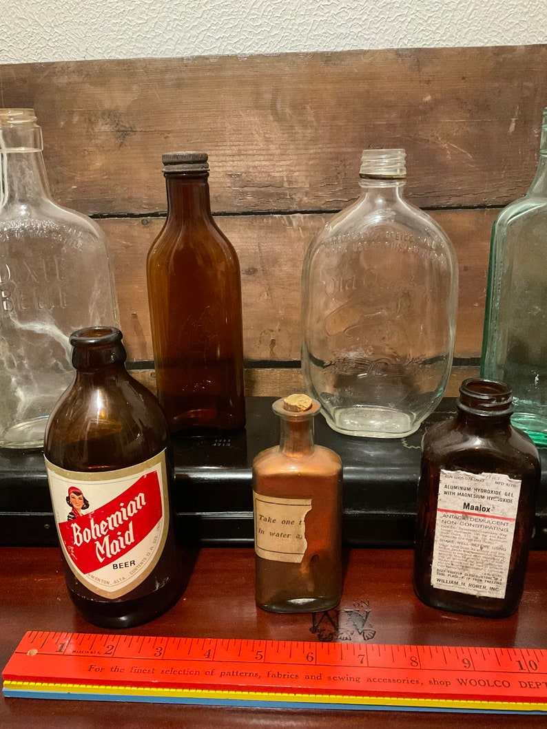 May include: A collection of vintage glass bottles, including a clear glass bottle with a cork stopper, a brown glass bottle with a label that reads "Bohemian Maid Beer", a clear glass bottle with a label that reads "Take one teaspoon in water", a brown glass bottle with a label that reads "Maalox Antacid Demulcent Non-Constipating", and a green glass bottle.