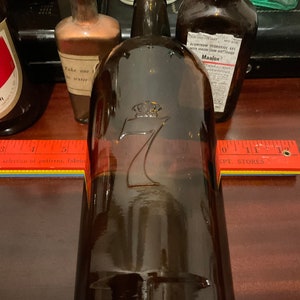 May include: A brown glass bottle with a crown and the number 7 embossed on the front. The bottle is sitting on a wooden surface with a ruler and a book.