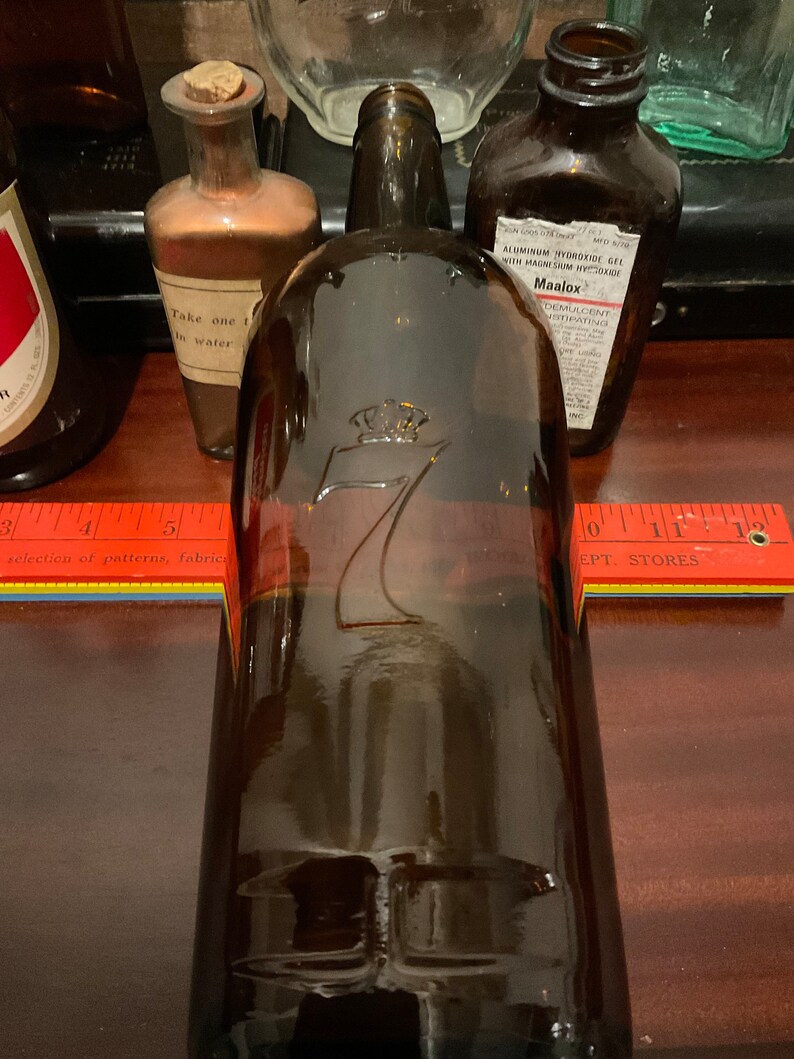 May include: A brown glass bottle with a crown and the number 7 embossed on the front. The bottle is sitting on a wooden surface with a ruler and a book.