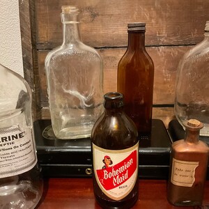 May include: A collection of vintage glass bottles, including a clear glass bottle with the text "Dixie Belle" on it, a brown glass bottle with a label that reads "Bohemian Maid Beer", and a clear glass bottle with a label that reads "Take one in water".