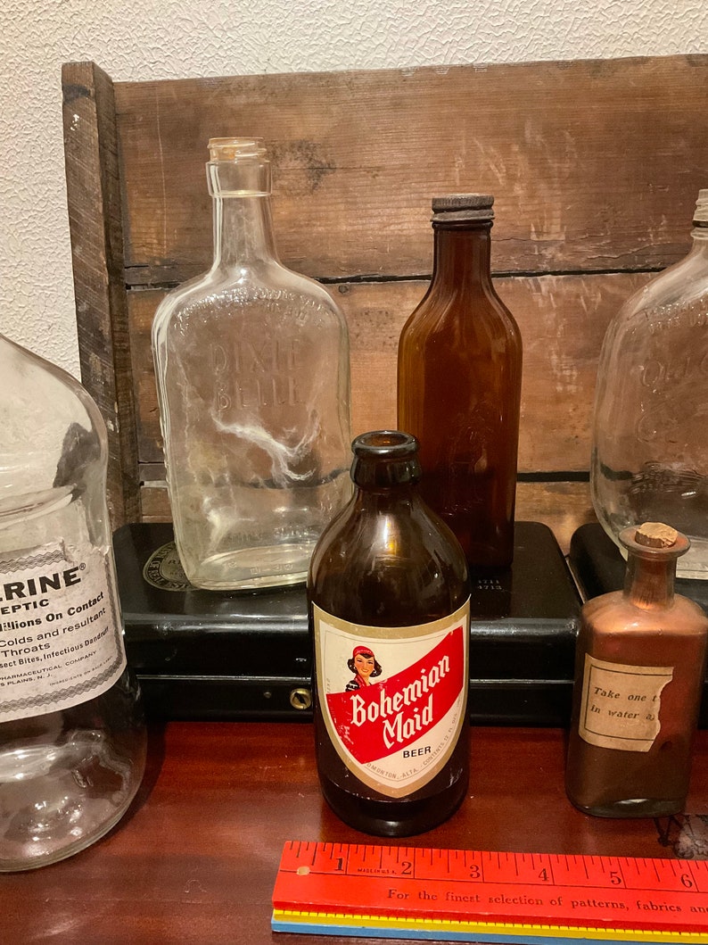 May include: A collection of vintage glass bottles, including a clear glass bottle with the text "Dixie Belle" on it, a brown glass bottle with a label that reads "Bohemian Maid Beer", and a clear glass bottle with a label that reads "Take one in water".