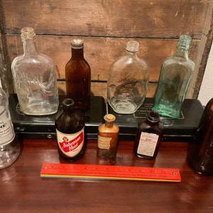 May include: A collection of vintage glass bottles, including a clear bottle labeled "Listerine", a brown bottle labeled "Bohemian Maid", and a green bottle labeled "Jack Daniels".