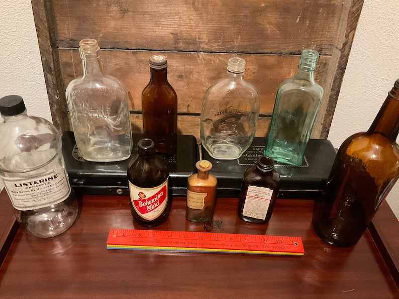 May include: A collection of vintage glass bottles, including a clear bottle labeled "Listerine", a brown bottle labeled "Bohemian Maid", and a green bottle labeled "Jack Daniels".