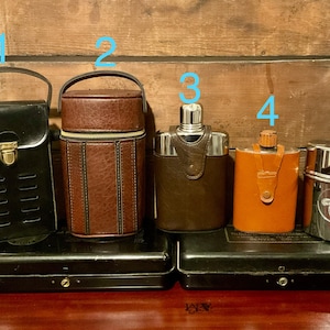 May include: A selection of vintage flasks. Features a black leather case, a brown leather flask with a zip, and three stainless steel flasks with leather or orange leather detailing. Various sizes and designs.
