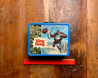 Op de afbeelding: Een rechthoekige blauwe metalen lunchtrommel met een afbeelding van King Kong op een wolkenkrabber, helikopters en de tekst King Kong. Een rode liniaal ligt onder de trommel en toont een breedte van ongeveer 23 cm voor het item.