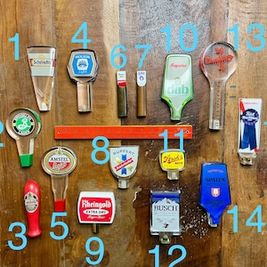 May include: A collection of fifteen beer tap handles featuring various brand logos including Schmidt's, Molson, Brooklyn Lager, Amstel Light, Ruppert, Stroh's, Busch, Spaten, and Pabst Blue Ribbon, arranged with an orange 12-inch ruler in the center.