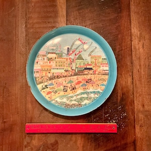 May include: A round, light blue metal tray with a detailed illustration of a beach scene. The illustration includes a Ferris wheel, roller coaster, beach umbrellas, and people. The tray measures approximately 12 inches in diameter.