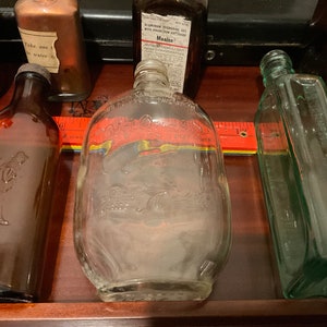 May include: Collection of vintage glass bottles, including a clear bottle with a raised design of a man and a woman, a clear bottle with a raised design of a bird, a brown bottle with a raised design of a man, and a green bottle with a raised design of a flower.