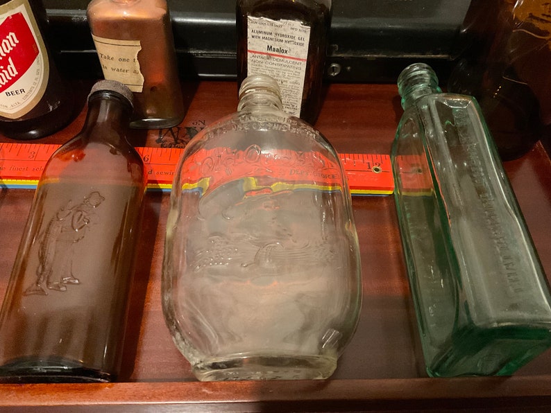 May include: Collection of vintage glass bottles, including a clear bottle with a raised design of a man and a woman, a clear bottle with a raised design of a bird, a brown bottle with a raised design of a man, and a green bottle with a raised design of a flower.