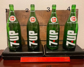 May include: Four vintage green glass 7 Up bottles with red and white labels. The bottles are 32 fluid ounces (1 quart) and have the text "Easy Open" and "Return for Deposit" on the labels. The bottles are sitting on black trays and a red ruler is in the foreground.