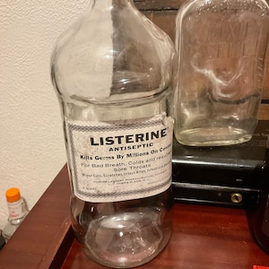 May include: A clear glass bottle with a black cap and a white label that reads "LISTERINE ANTISEPTIC Kills Germs By Millions On Contact For Bad Breath, Colds and resulting Sore Throats Minor Cuts, Scratches, Insect Bites, Infectious diseases WARNER-LAMBERT PHARMACEUTICAL COMPANY 1 QUART MORRIS PLAINS, N.J."