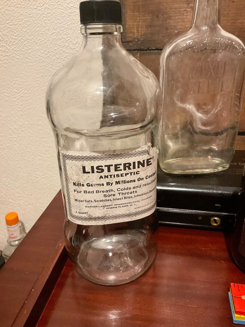 May include: A clear glass bottle with a black cap and a white label that reads "LISTERINE ANTISEPTIC Kills Germs By Millions On Contact For Bad Breath, Colds and resulting Sore Throats Minor Cuts, Scratches, Insect Bites, Infectious diseases WARNER-LAMBERT PHARMACEUTICAL COMPANY 1 QUART MORRIS PLAINS, N.J."