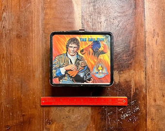 Op de afbeelding: Een rechthoekige metalen lunchtrommel met een The Fall Guy-afbeelding van een man die een helm vasthoudt tegen een vurige oranje achtergrond. De tekst THE FALL GUY staat in blauw gedrukt. Een rode liniaal eronder toont 23 cm.