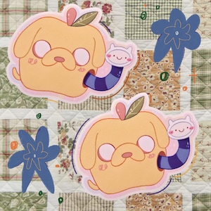 May include: Two cartoon stickers featuring a yellow dog-like character with a pink nose and ears, and a small white creature with a blue and purple striped tube. The stickers are on a patchwork quilt background with blue flower shapes.