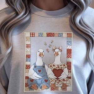 Könnte beinhalten: Hellgraues Sweatshirt mit einem gerahmten Motiv von zwei Gänsen. Eine Gans trägt blaue Latzhosen mit Herzdetails, die andere ein Blumenkleid und hält ein Herz. Der Rahmen hat ein Patchwork-Design.