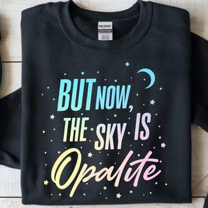 May include: Black crewneck sweatshirt with the text "BUT NOW, THE SKY IS Opalite" in a colorful, celestial design. The design includes stars and a crescent moon. The sleeves are cuffed.
