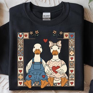 May include: Black sweatshirt featuring a charming illustration of two ducks in love. One duck wears overalls, the other a dress, both holding hearts. The design is framed with hearts and floral accents, perfect for Valentine's Day.